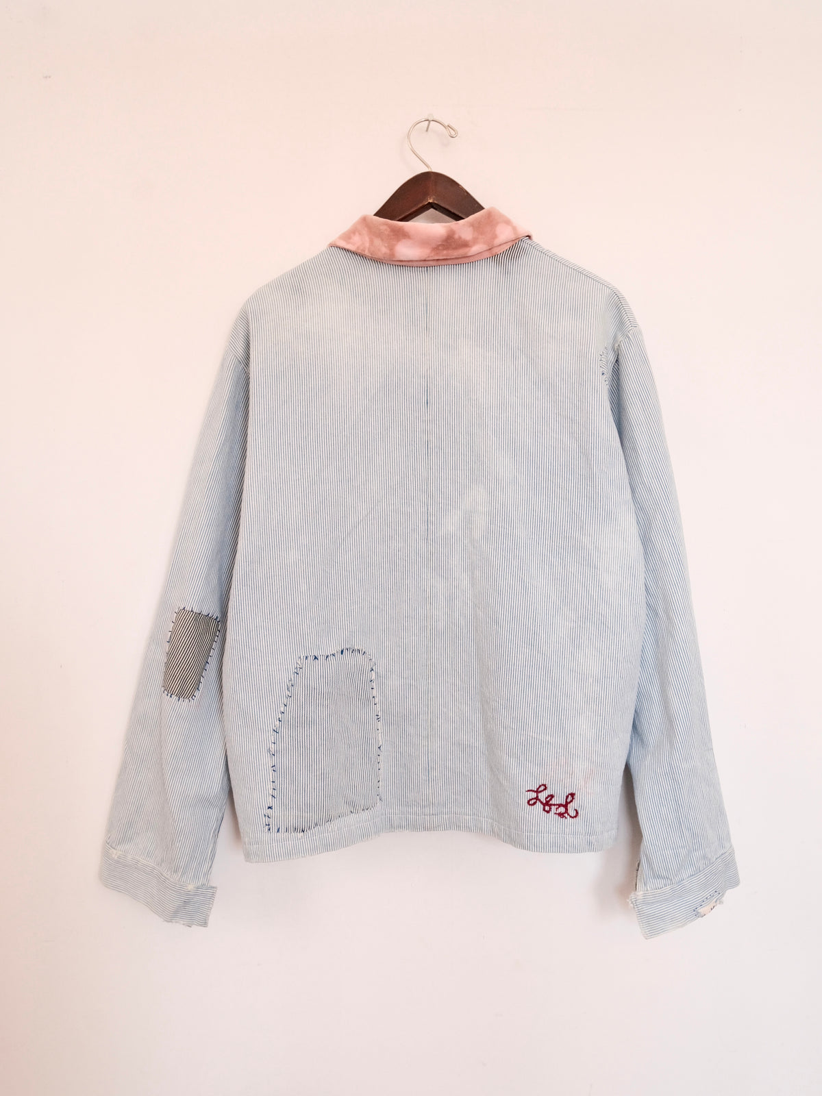 A light blue denim jacket hanging on a wooden hanger against a plain off-white wall. The back of the jacket features a pink satin collar, a gray fabric patch on one sleeve, a large raw-edge patch pocket on the lower back, and red embroidered initials 'LS' on the lower right.