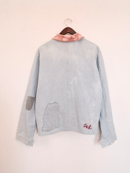 A light blue denim jacket hanging on a wooden hanger against a plain off-white wall. The back of the jacket features a pink satin collar, a gray fabric patch on one sleeve, a large raw-edge patch pocket on the lower back, and red embroidered initials 'LS' on the lower right.