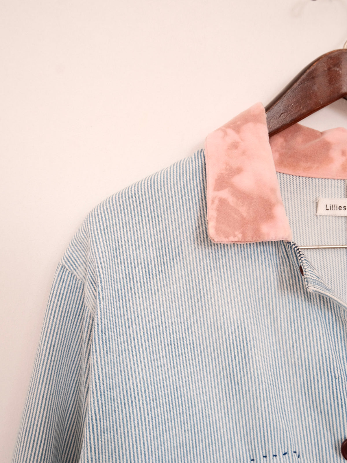 A close-up of a light blue and white vertically striped button-up shirt hanging on a wooden hanger against a plain off-white wall. The shirt features a contrasting dusty pink velvet collar, and a brand label reading 'Lillies' is visible inside the collar.