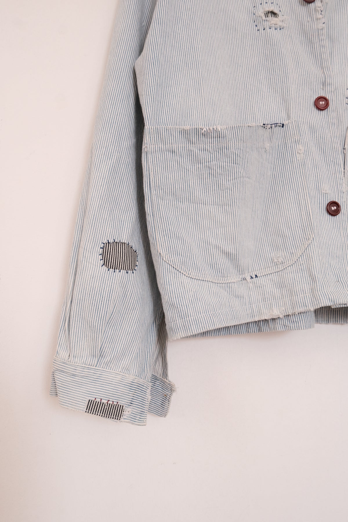 Close-up of a light blue and white striped distressed denim jacket, showing a front pocket, brown buttons, and ripped details on the chest and sleeve.