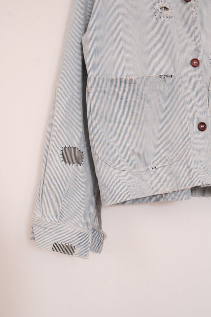 Close-up of a light blue and white striped distressed denim jacket, showing a front pocket, brown buttons, and ripped details on the chest and sleeve.