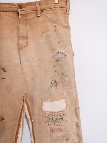 Distressed tan denim jeans with heavy paint splatter, ripped details, and a pink 'LIVE' embroidery on the lower leg, shown against a plain white background.